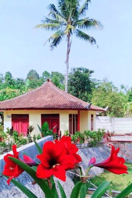 A charming traditional house features a distinctive thatched roof and red wooden doors. Surrounding greenery adds a natural touch, complemented by vibrant red flowers in the foreground. A tall palm tree rises beside the structure, enhancing the tranquil setting.
