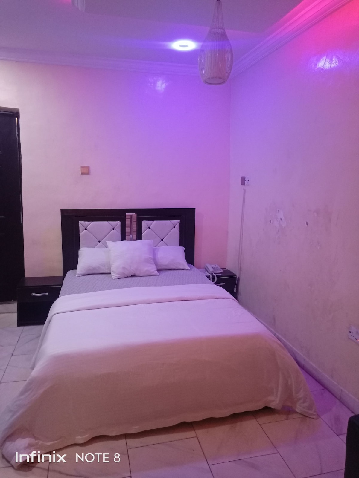 A compact sleeping area features a snug bed adorned with white linens and multiple white pillows. The minimalist design includes bedside tables, with a soft glow emanating from a ceiling light that casts a gentle hue on the pastel-colored walls.