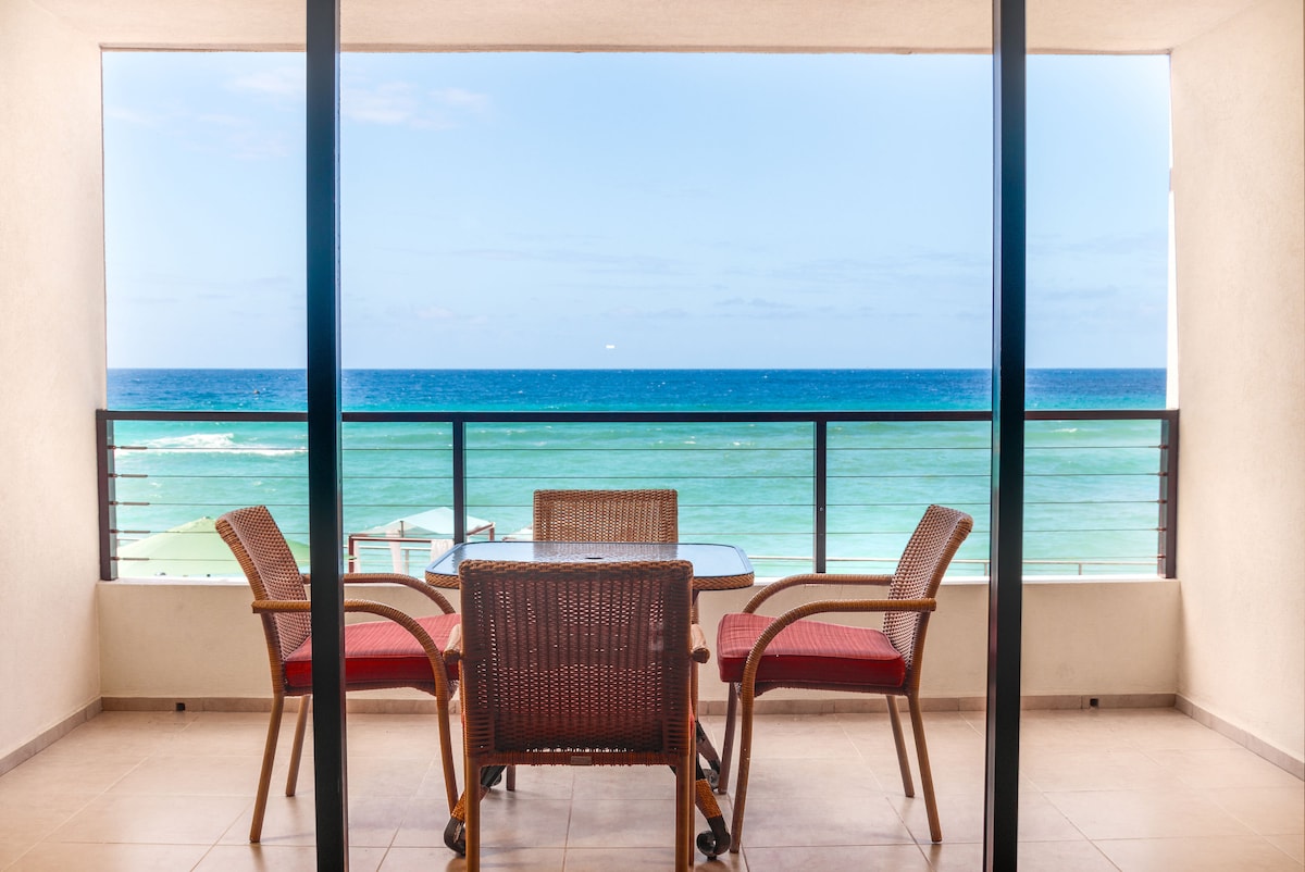 A private terrace offers a clear view of the blue Caribbean sea, framed by two glass doors. A table with four cushioned chairs stands in the foreground, inviting relaxed outdoor dining. The horizon features gentle waves and a bright sky, enhancing the serene atmosphere.