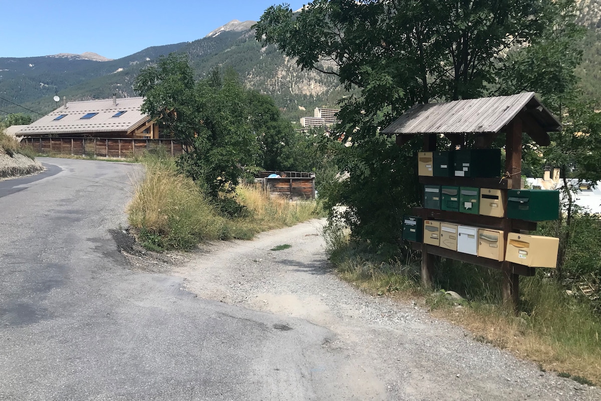A gravel path diverges towards a wooded area, with clusters of trees providing shade. A wooden structure stands nearby, displaying several mailboxes of varying sizes and colors. In the background, a building featuring solar panels is visible against a mountainous backdrop.