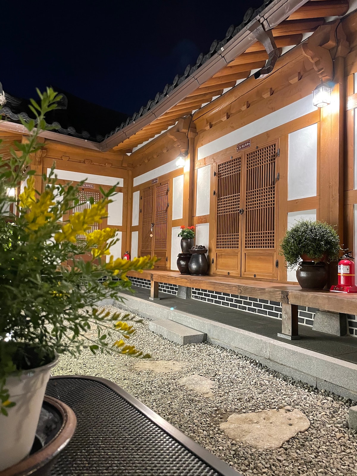 The traditional hanok structure is framed by wooden eaves and sliding doors, emanating a warm glow from outdoor lighting. A stone pathway leads to a garden area featuring potted plants and decorative stones, enhancing the tranquil ambiance of the space.