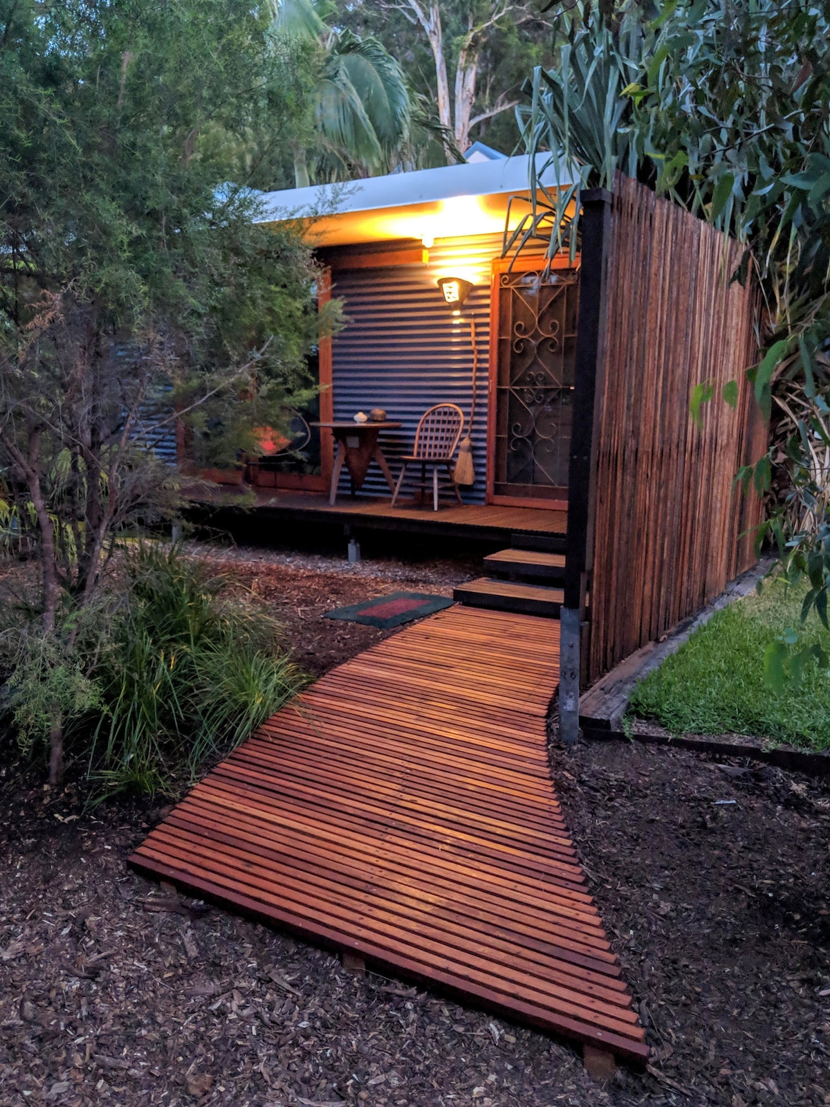 A quaint eco retreat is set within a garden, featuring a wooden pathway leading to a private entrance. Soft lighting illuminates the exterior, while outdoor seating is positioned on a small patio area, surrounded by natural greenery and trees.