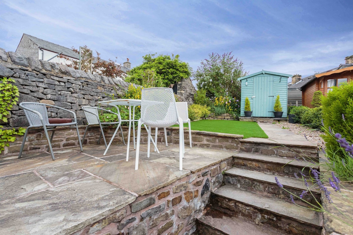 A spacious outdoor patio area features a set of white metal chairs arranged around a small table. Stone steps lead down to the garden, which includes a well-maintained green lawn and a blue garden shed in the background, framed by a low stone wall.