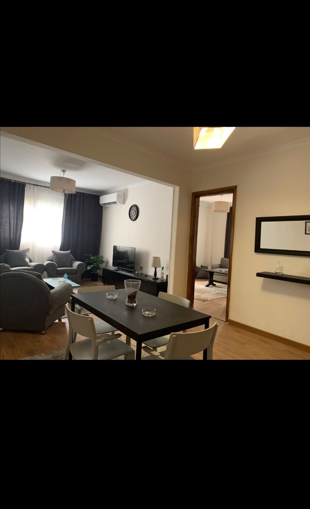 The living area features a comfortable seating arrangement with soft couches and a television. A dining table is positioned adjacent, accompanied by four chairs. Natural light filters in through large windows, complementing the modern decor and inviting atmosphere.