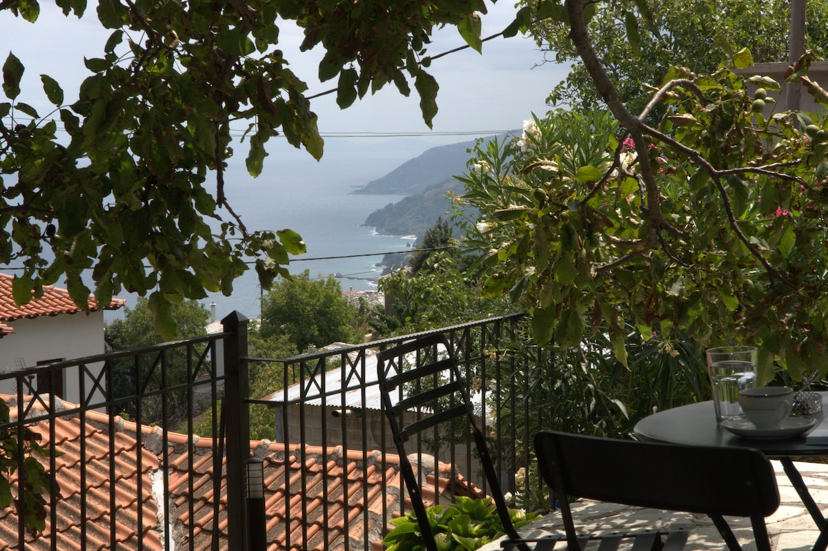 A view from a balcony framed by lush greenery overlooks the sea and distant mountains. A small black table with chairs is positioned on the terrace, set against a backdrop of traditional stone rooftops, highlighting a serene and inviting outdoor space.