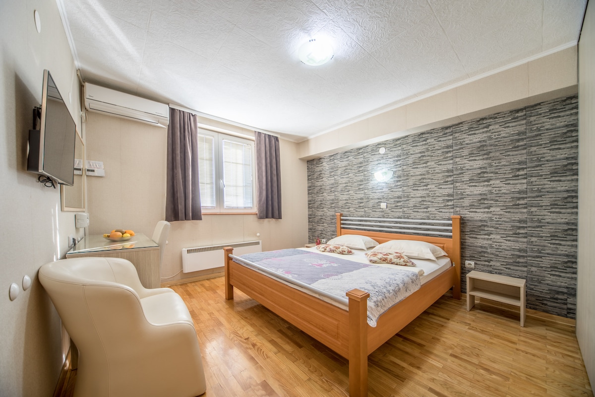 A bright bedroom features a wooden bed with decorative bedding and a textured accent wall. A small desk and chair are positioned near a window, while a flat-screen TV is mounted on the opposite wall. Natural light filters through light curtains.