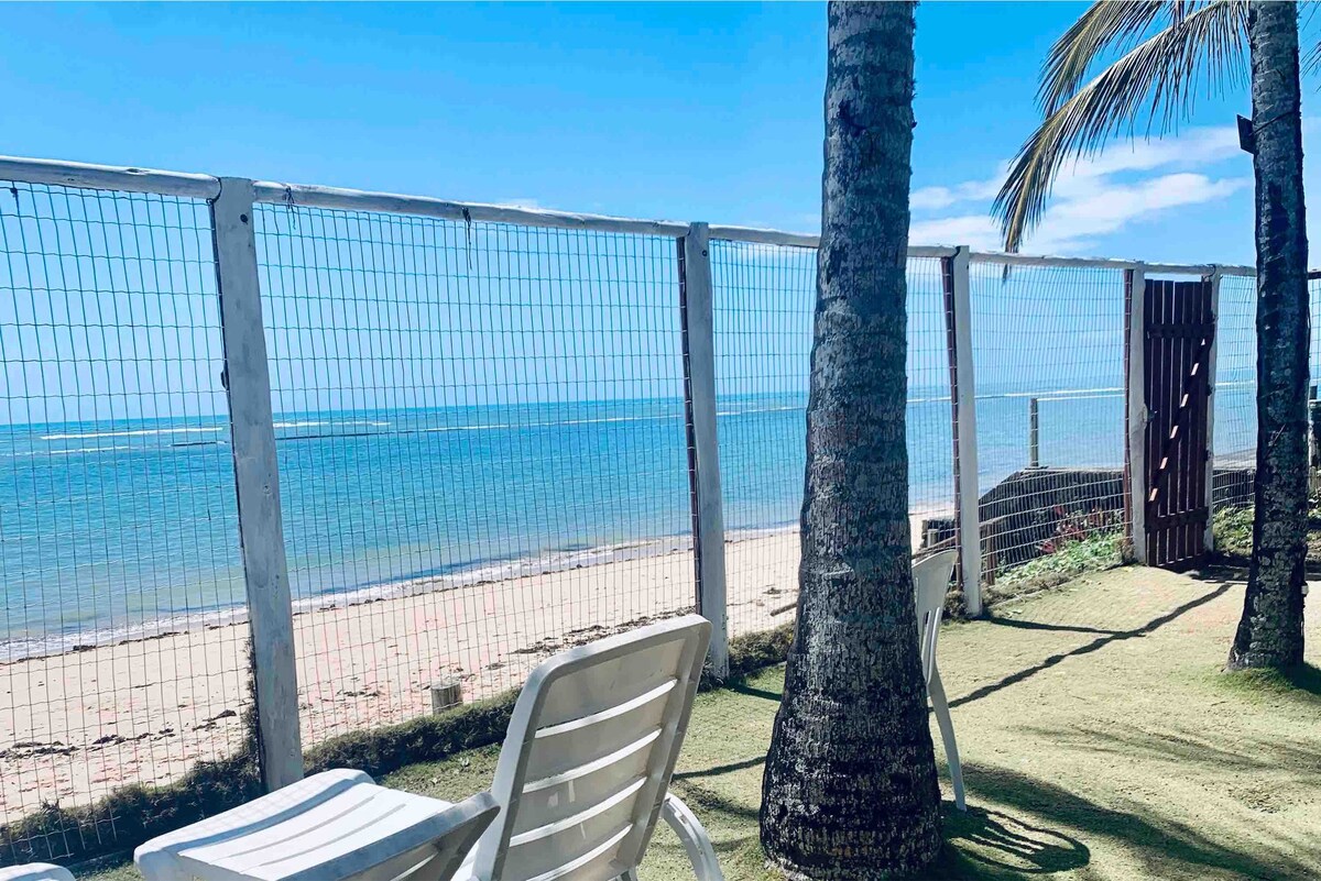 A serene beachfront view is captured, featuring a sandy shoreline meeting calm blue waters. Two white lounge chairs are positioned under palm trees, providing a relaxing spot. A fence runs along the area, ensuring privacy while still offering unobstructed views of the horizon.