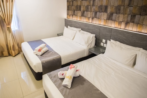 A well-appointed guest room is featured, containing two spacious beds with crisp white linens. A textured accent wall adds interest, illuminated by soft lighting. Fresh towels are neatly arranged on the beds, and natural light filters in through a nearby window draped with elegant curtains.
