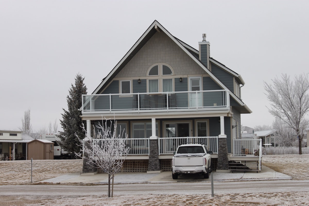 A modern two-story house features a large front balcony and a spacious porch, adorned with a railing. A white vehicle is parked on the driveway. Surrounding trees are frosted, and the landscape is coated with a light layer of snow, contributing to a tranquil winter scene.