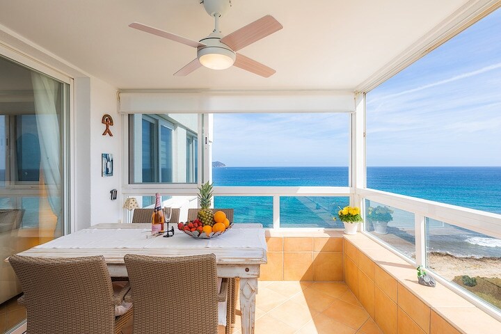 A sunlit dining area features a wooden table set with fresh fruit and beverages, overlooking the sea through large panoramic windows. Light-colored wicker chairs surround the table, and a potted plant adds a touch of greenery to the space.
