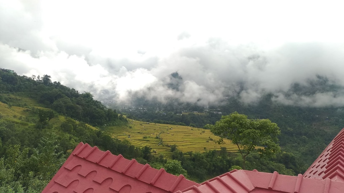 A scenic view illustrates lush terraced fields beneath a cloudy sky. Green hills rise in the background, with patches of gold from ripe crops visible in the fields. The red roof of the cottage partially frames the image, offering a glimpse of the natural landscape.