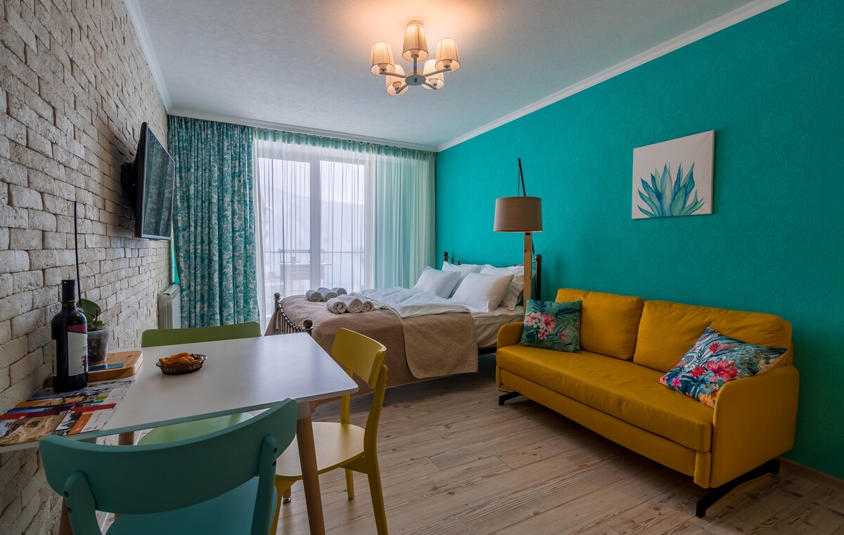 A vibrant living space is presented with an azure accent wall and light wood flooring. A spacious bed is arranged alongside a cheerful yellow sofa. A small dining table with colorful chairs is visible, along with a decorative piece on the wall and a view through large windows.