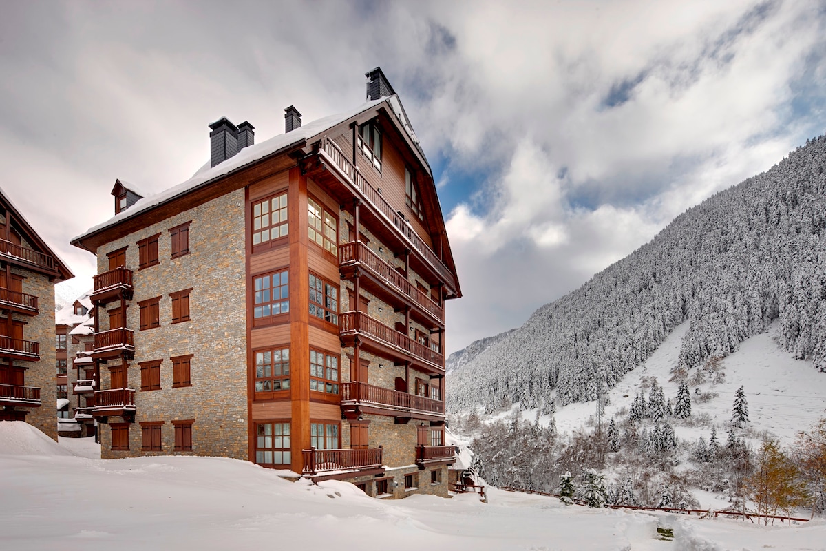 The exterior of a multi-story building is displayed, featuring a blend of stone and wood accents. Balconies are visible on each level, offering views of the surrounding snow-covered landscape. Rolling hills and tree-covered slopes are seen in the background under a partly cloudy sky.