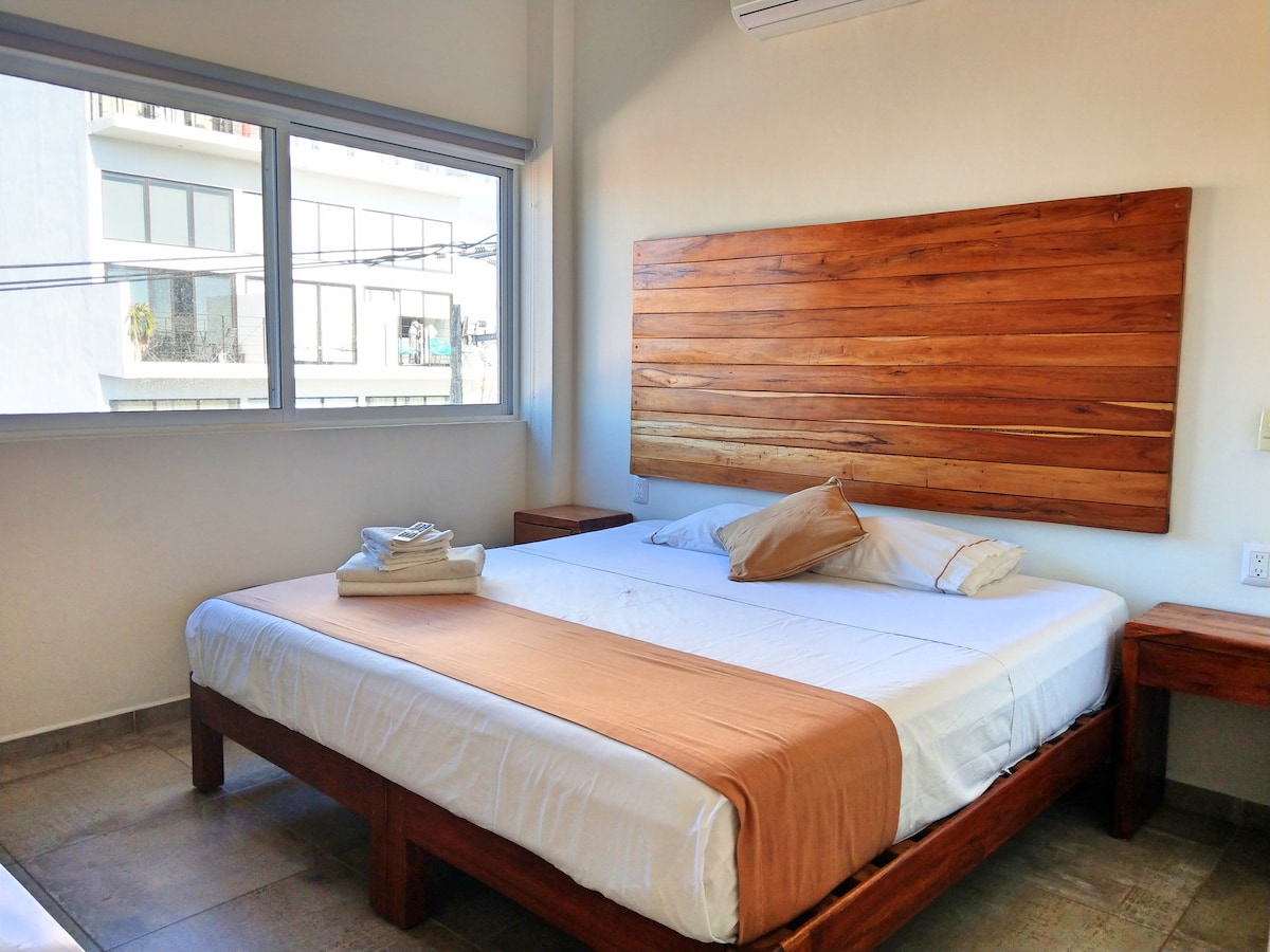 A simple bedroom features a spacious bed with a wooden headboard. Two nightstands are positioned on either side of the bed. Natural light fills the room through a large window, offering views of the outside. Crisp white linens and neatly arranged towels can be seen.