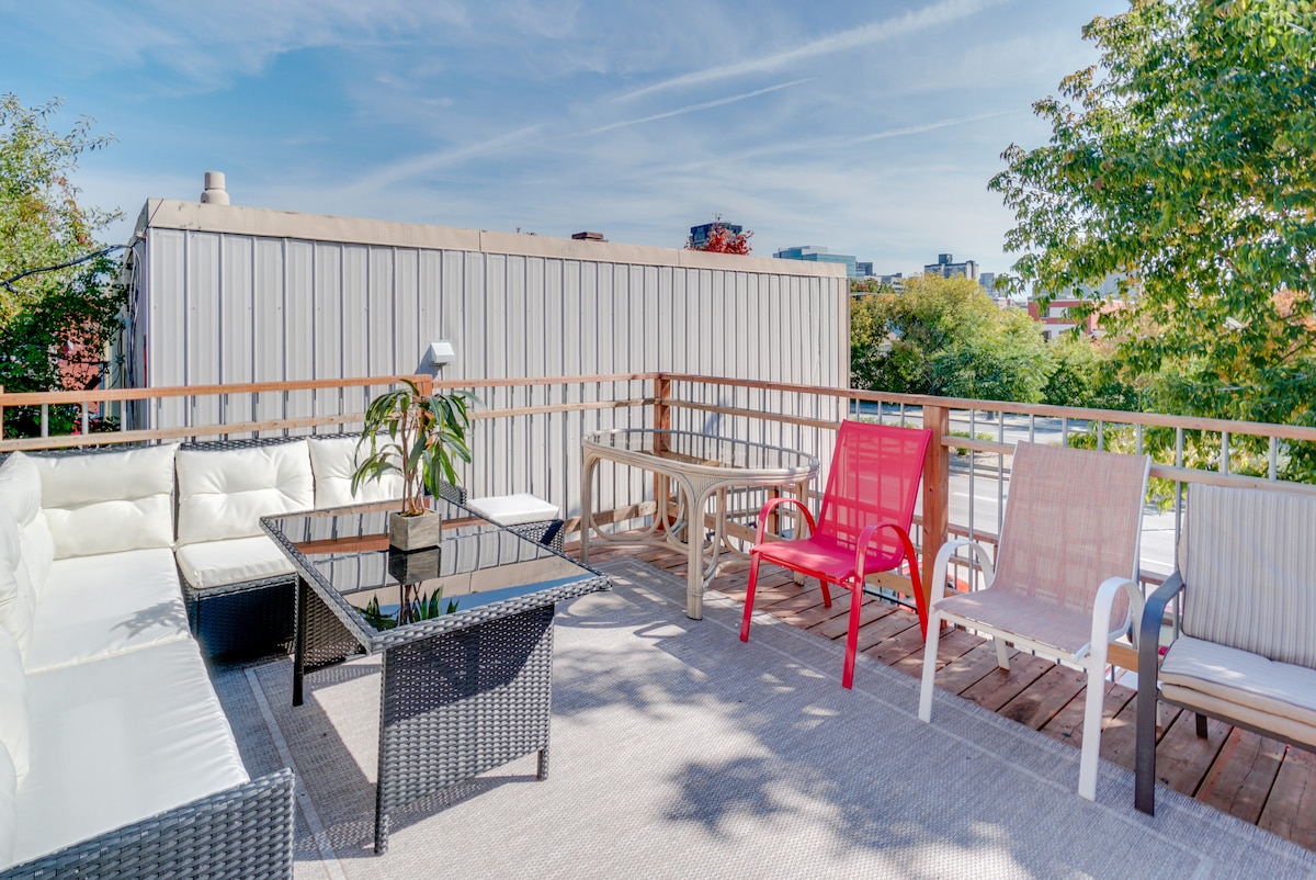 The rooftop patio is furnished with a variety of seating options, including a white sectional sofa and metal chairs in red and white. A glass-topped table is situated in the center, surrounded by wood decking and greenery in the background, with clear skies overhead.