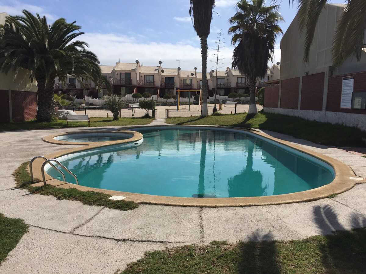 A tranquil pool area is surrounded by lush palm trees and a well-maintained lawn. The pool features a unique shape with a shallow section, inviting relaxation. Nearby, the condominium's common spaces are visible, offering an open environment for leisure and socializing.