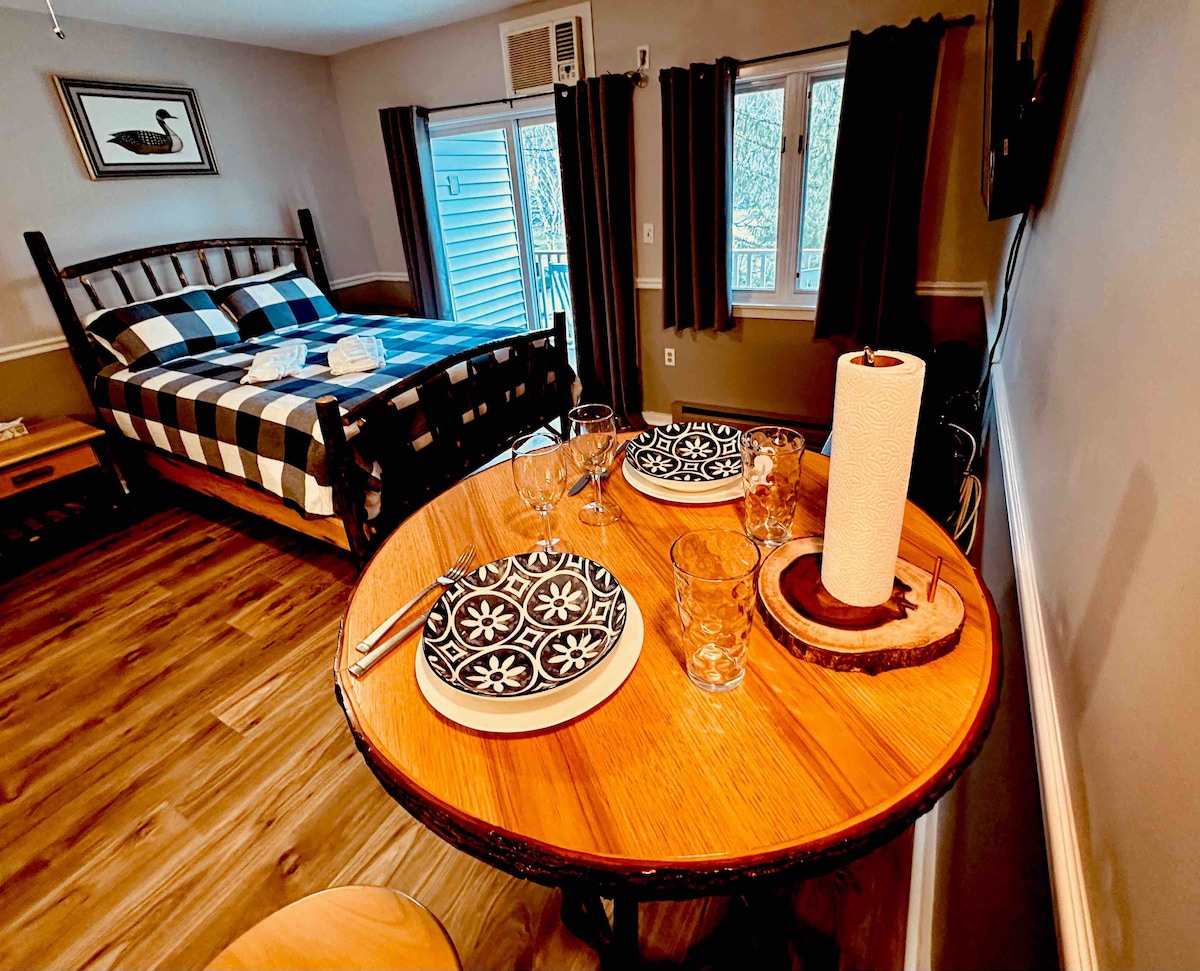 A cozy studio features a dining table set with two decorative plates and glasses, adjacent to a spacious bed dressed in a checked blanket. Natural light is welcomed by the nearby window, and dark curtains frame the view, enhancing the inviting atmosphere of the space.