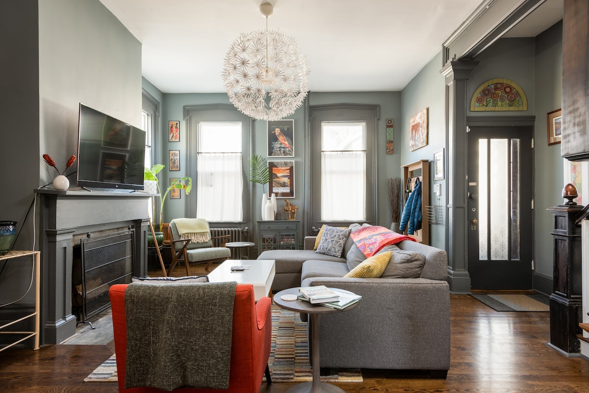 A cozy living room features a large sectional sofa in neutral tones, positioned near a decorative fireplace. Natural light enters through two windows, creating a bright atmosphere. A modern light fixture hangs overhead, while various pieces of artwork adorn the walls, adding character to the space.