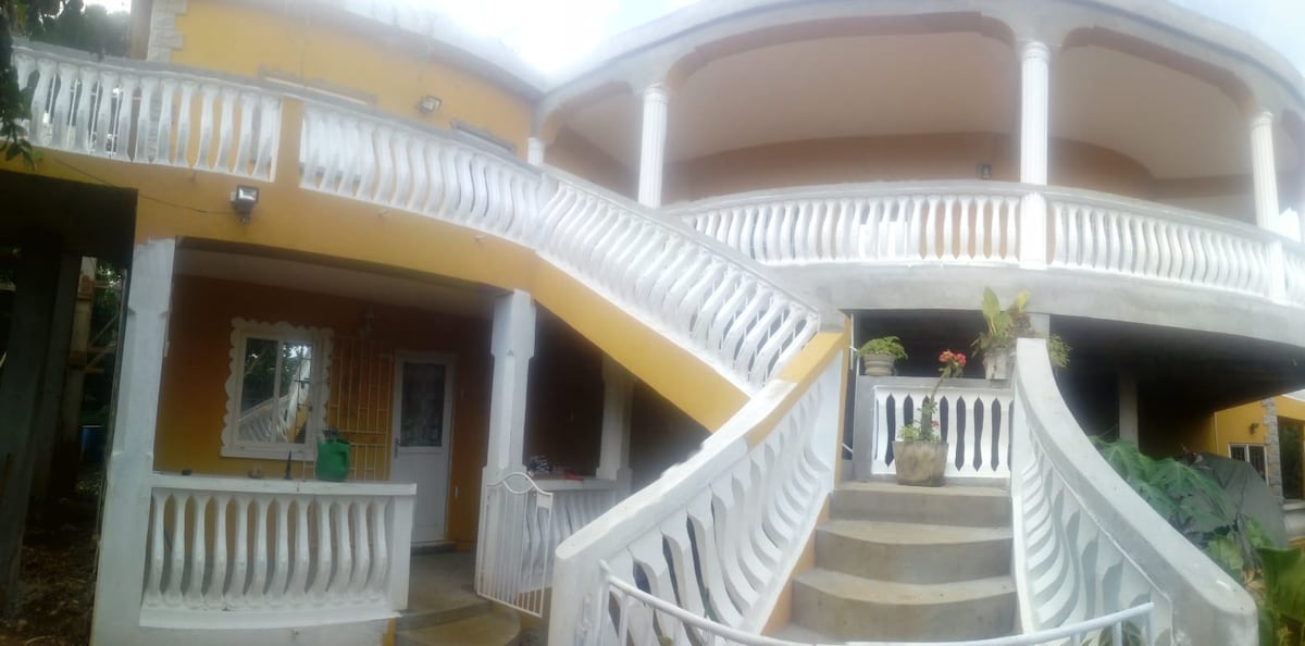 A two-level property is displayed, featuring a large staircase with decorative railings leading to upper balconies. The exterior is painted in warm tones, and tropical plants can be seen along the entrance. The inviting architecture promotes an open and airy feel.
