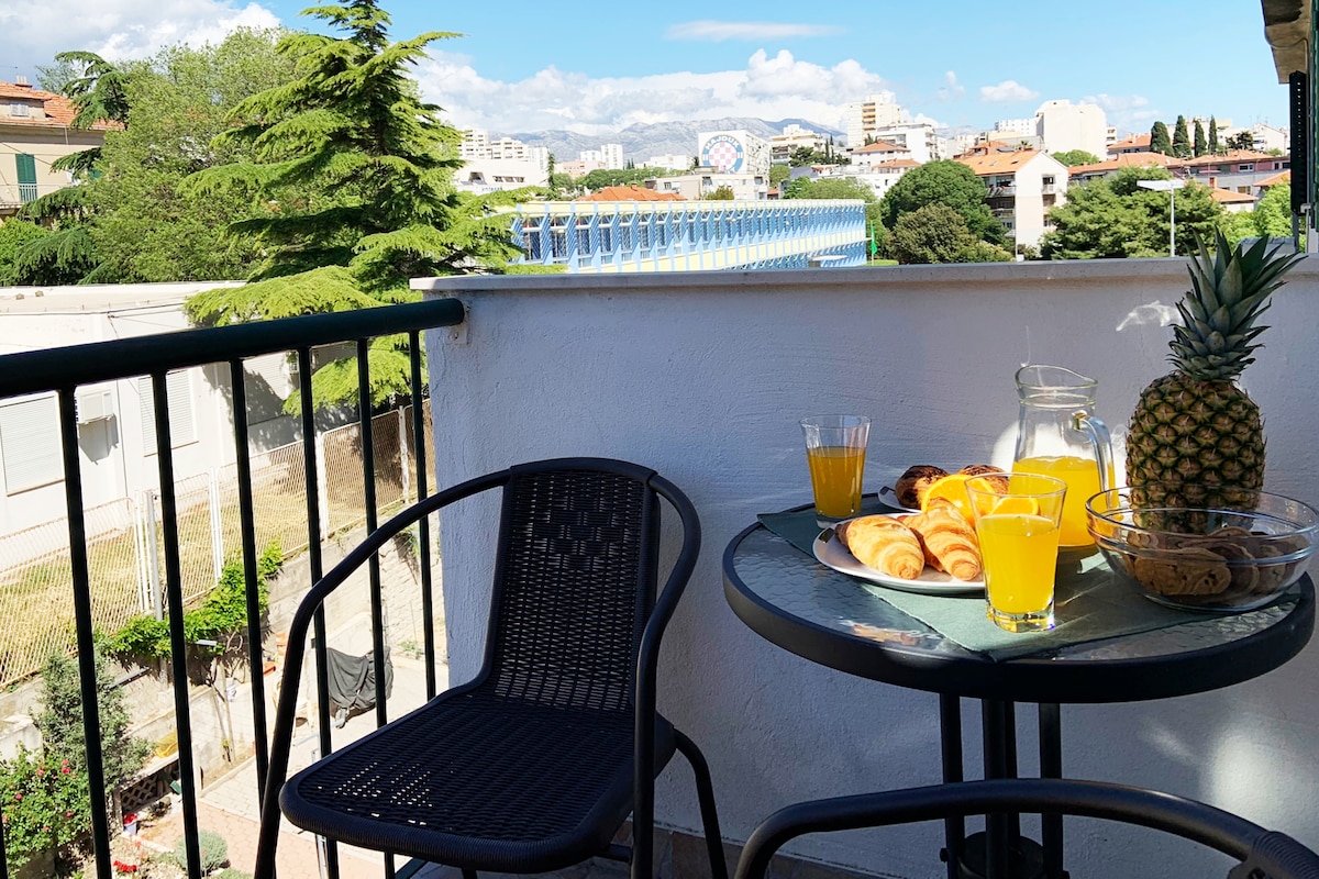 A private balcony is shown, featuring a small black table with a pitcher and glasses filled with orange juice. Fresh pastries and cookies are arranged on a green serving tray. Two black chairs are positioned beside the table, surrounded by greenery and city views in the background.