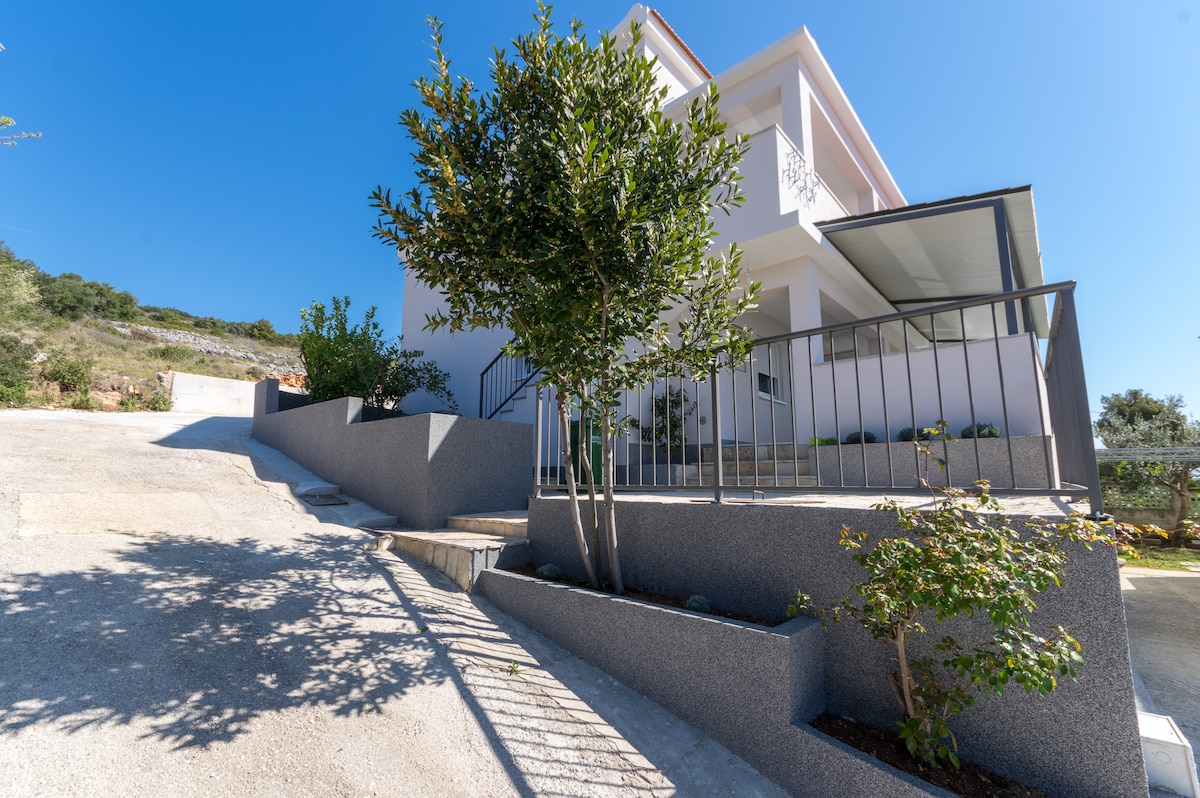 The exterior of the apartment showcases a modern design with light-colored walls and a covered entrance. Lush greenery and planters adorn the pathway, creating a welcoming approach. The sloped driveway and scenic hillside are visible in the background, highlighting the peaceful surroundings.