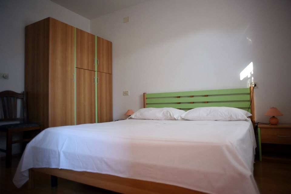 A bright sleeping area features a spacious bed with white linens. A wooden wardrobe with green accents stands against the wall. Soft lighting from bedside lamps creates a calm atmosphere, while a simple wooden chair is positioned nearby.