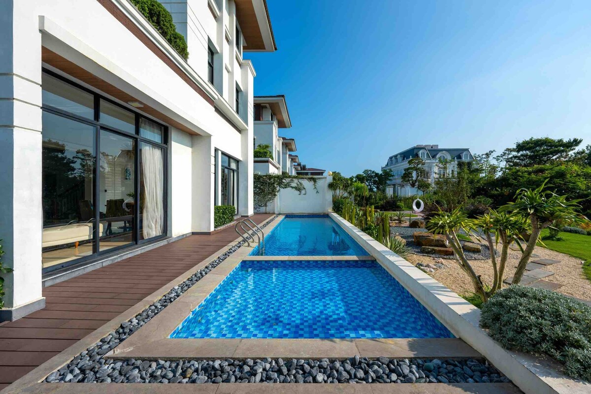 A private pool stretches alongside the villa, featuring shimmering blue tiles. Lush greenery and landscaped areas are visible, framed by the villa's modern architecture. Sunlight reflects off the pool's surface, enhancing the serene outdoor setting.