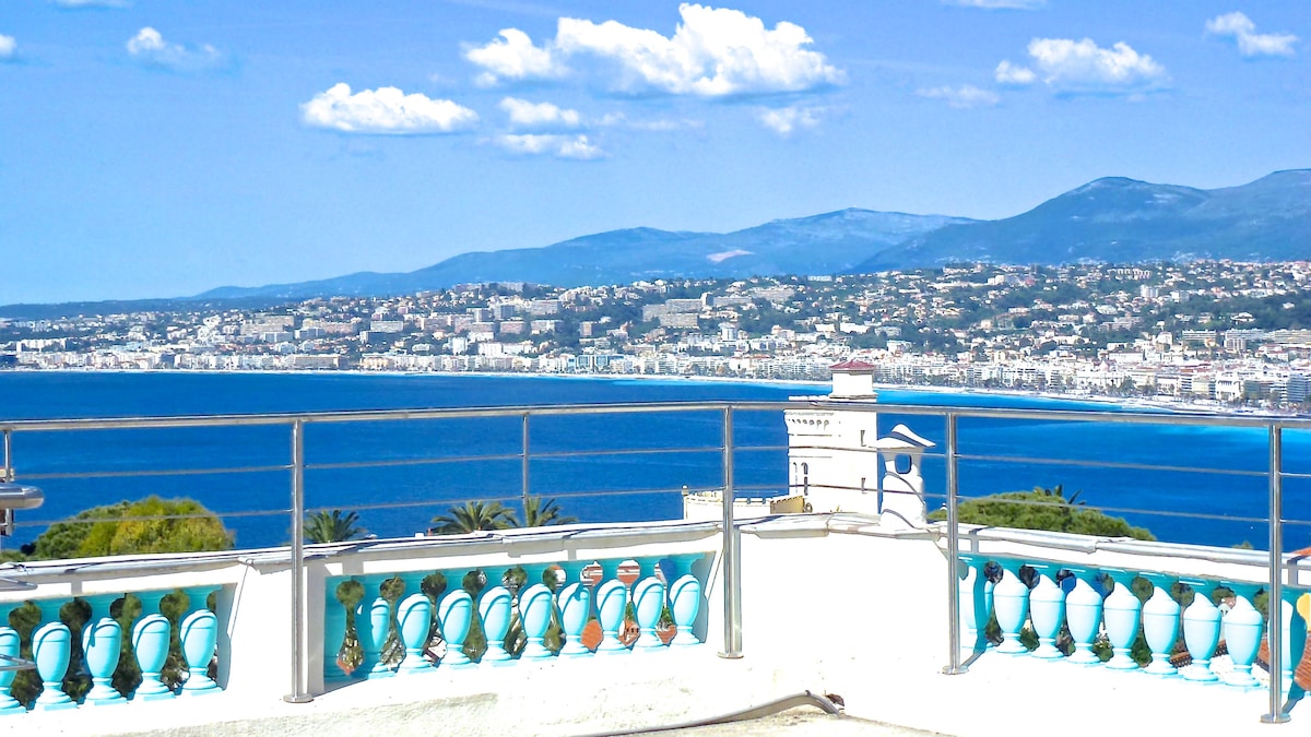 A panoramic view of the Mediterranean coastline is displayed from a terrace. The azure waters reflect sunlight, complemented by the vibrant blue sky. The landscape features the city of Nice and distant mountains, with a railing adorned with turquoise accents enhancing the scenic outlook.