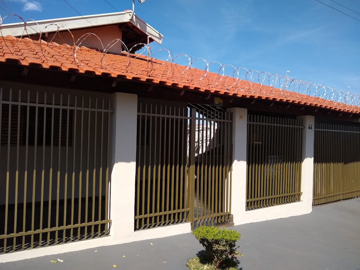 The exterior of the property features a decorative barbed wire fence atop a sturdy metal gate. The entrance offers clear access to the front of the house. The building's roof is made of terracotta tiles, contrasting with the beige walls and lush greenery nearby.