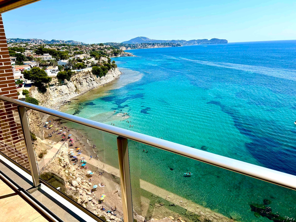 A balcony view presents a sweeping vista of the coastline, highlighting tranquil turquoise waters. The scene captures a sandy beach lined with small groups of sunbathers. Rolling hills and distant towns complete the picturesque landscape under clear blue skies.
