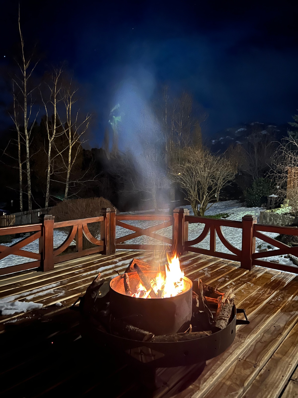 A circular fire pit sits on a wooden deck, with flames creating a warm glow against the dark night sky. Surrounding trees are silhouetted, and snow can be seen on the ground, enhancing the peaceful outdoor setting.