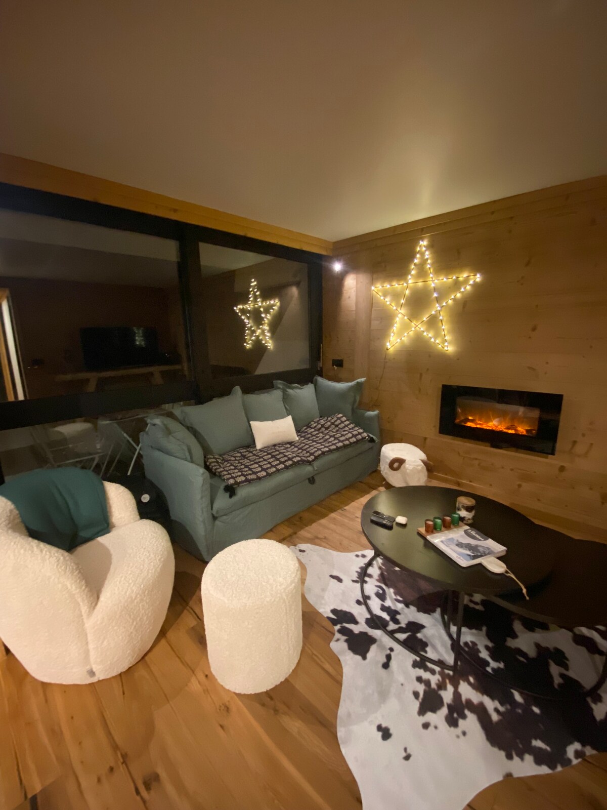 A comfortable living area features a plush sofa and cozy armchairs, illuminated by soft lighting. A decorative star made of lights is displayed on the wall, alongside an electric fireplace that adds warmth. A low coffee table showcases a few decorative items on a patterned rug.