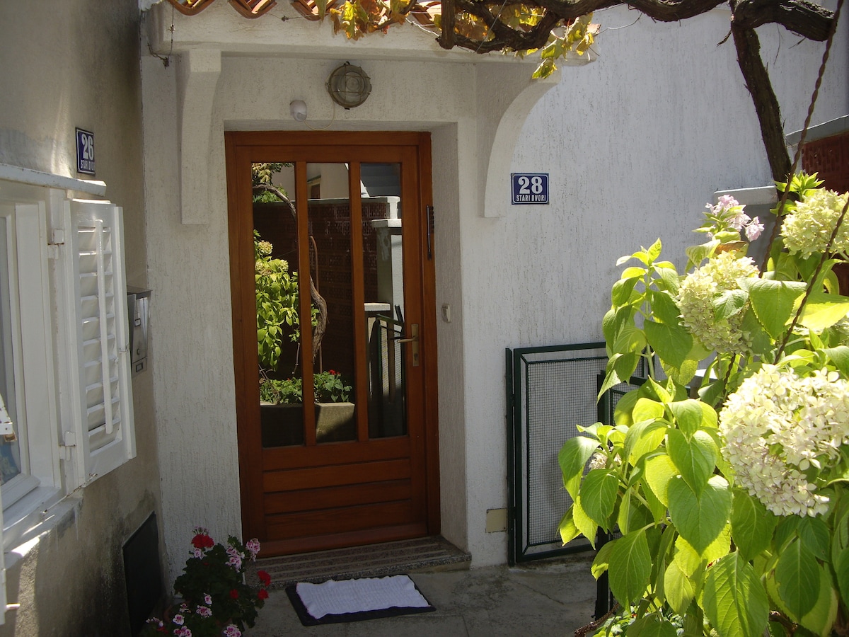 The entrance features a wooden door flanked by a lush garden, showcasing a variety of plants and flowers. A welcome mat lies in front of the door, and a decorative house number is displayed above the entryway.