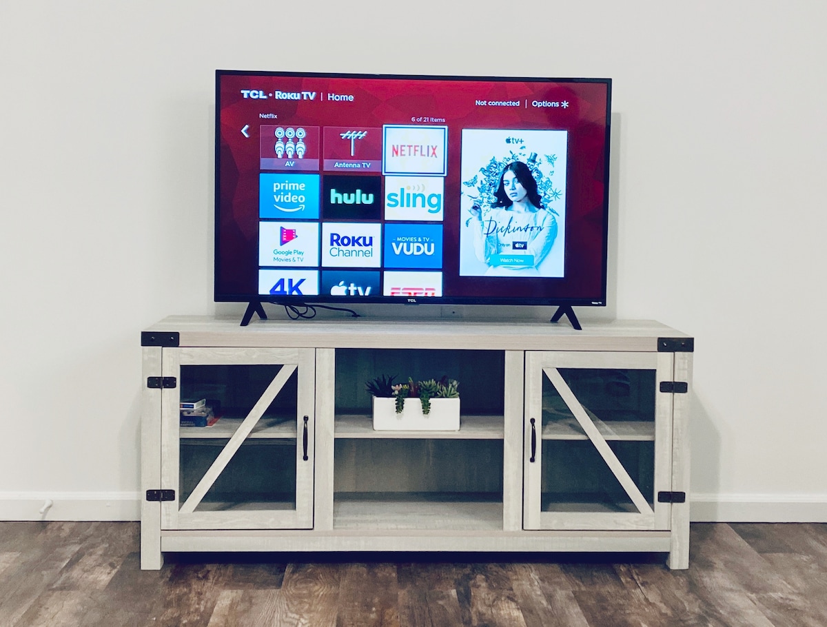A flat-screen TV is mounted on the wall, displaying various streaming service options. Below the TV, a light-colored wooden media console features glass cabinet doors and decorative items, including a small potted plant, enhancing the modern ambiance of the space.