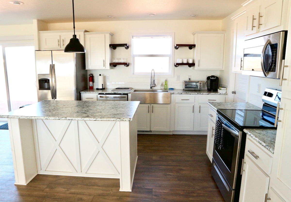 A modern kitchen features sleek white cabinetry with contrasting dark stainless steel appliances. A central island, topped with textured granite, offers additional preparation space. Natural light from a window brightens the area, which is arranged for convenience and functionality.