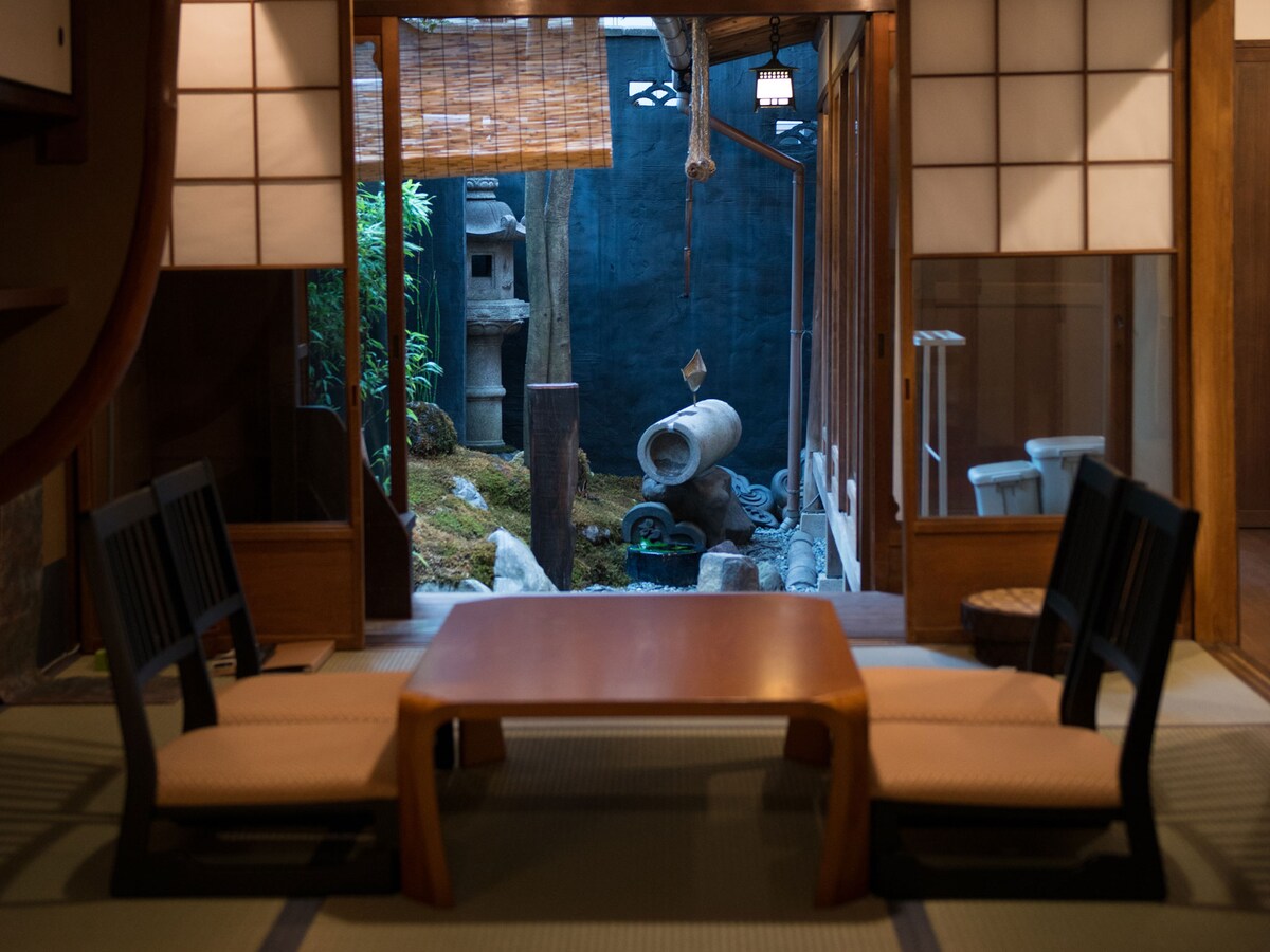 A traditional tatami room features a low wooden table surrounded by four black chairs. Large sliding doors provide a view of a serene garden with bamboo and stone elements. Natural light filters in, highlighting the calm atmosphere of the space.