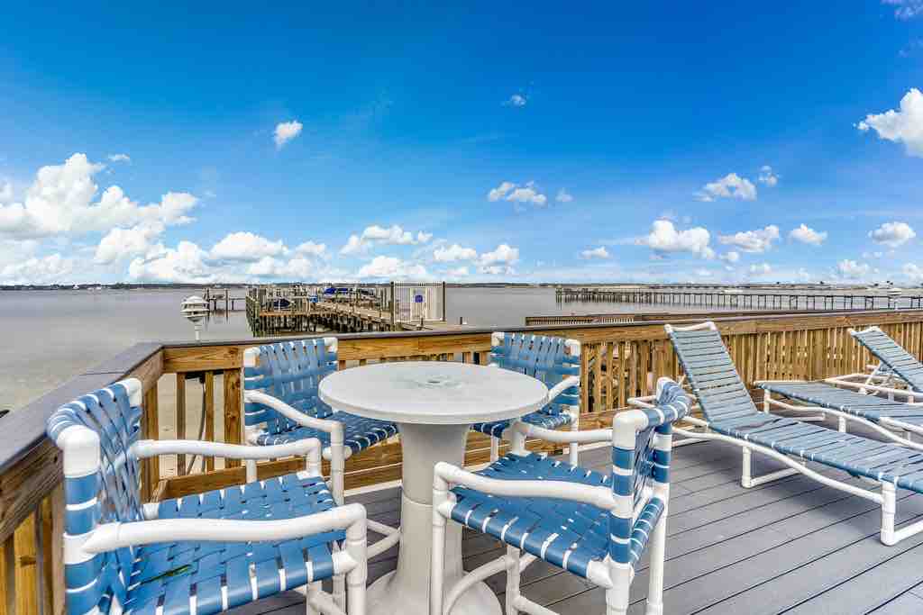 An outdoor deck features a round table surrounded by four blue and white chairs, complemented by two lounge chairs positioned for relaxation. The expansive view overlooks the water, with a pier visible in the background against a bright, sunny sky.