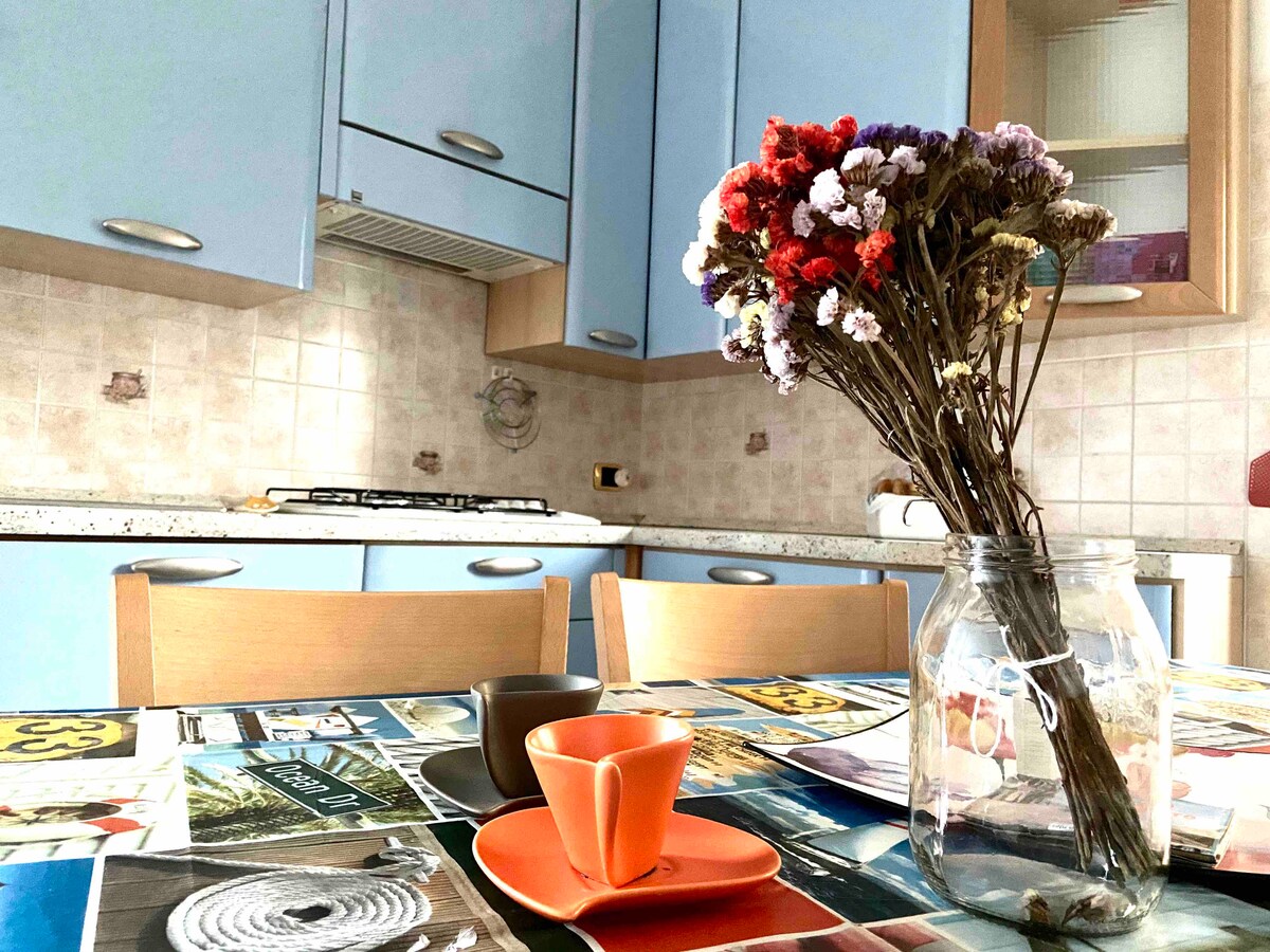 The kitchen area features light blue cabinetry and a countertop with a gas stove. A table is set with orange dishware and has a vase of dried flowers, adding a touch of color. The walls are tiled in a subtle pattern, enhancing the space's inviting feel.