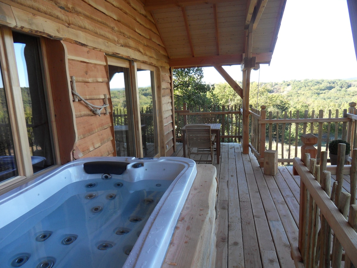A private terrace showcases a wooden deck with a hot tub, inviting relaxation. Nearby, a small dining table with chairs offers a serene view of the surrounding nature, while large windows enhance the connection to the outdoors.