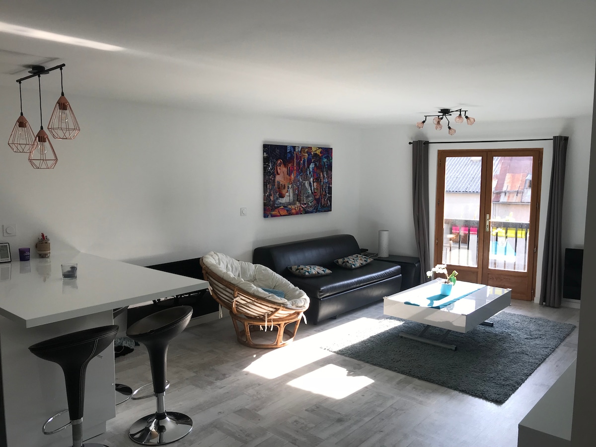 A modern living area features a spacious black sofa and a round wicker chair, complemented by a white coffee table atop a soft grey rug. Natural light floods through the large double doors leading to a charming balcony, with colorful artwork displayed on the white wall.