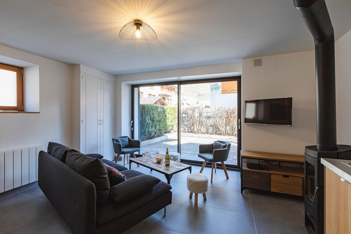 The living area features a neutral color palette and includes a spacious sofa, a wooden coffee table, and a small pouf. A large glass door opens to the outdoor terrace, allowing natural light to fill the room. A wall-mounted television and a wood stove are also visible.