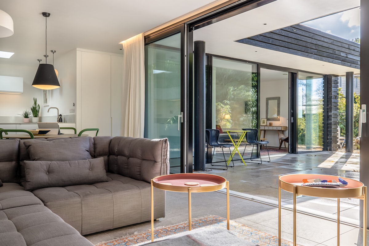 An open living area features a modern, plush sectional sofa facing a large glass wall that connects to the outdoor space. Two small, round tables, one with a decorative tray, are positioned in front. Natural light fills the room, highlighting the simple decor.