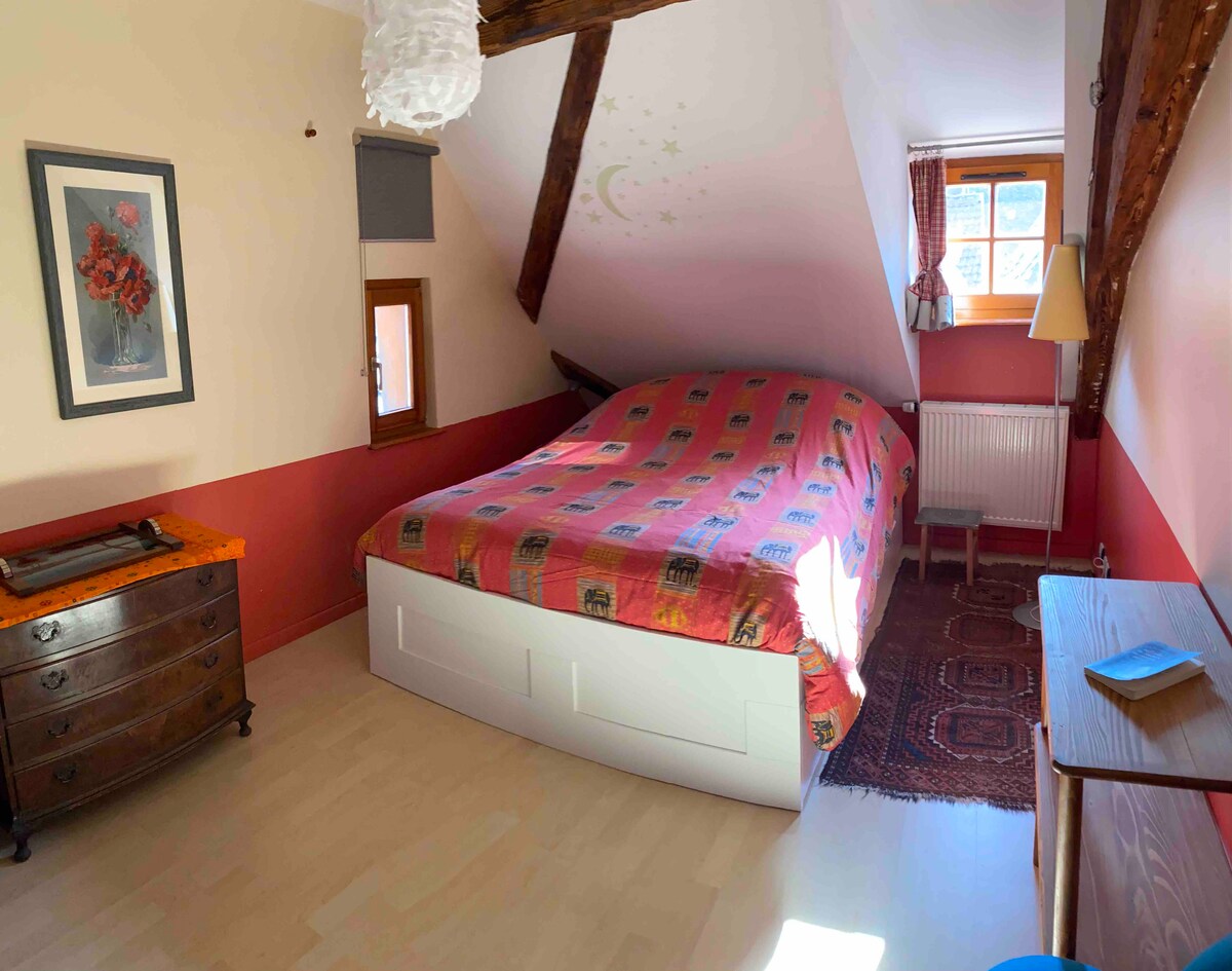 A charming attic bedroom features a spacious bed covered with a vibrant patterned duvet. Exposed wooden beams add character, while natural light filters in through a window, highlighting a tasteful blend of colors on the walls. A wooden dresser and a small table are present, creating a cozy atmosphere.