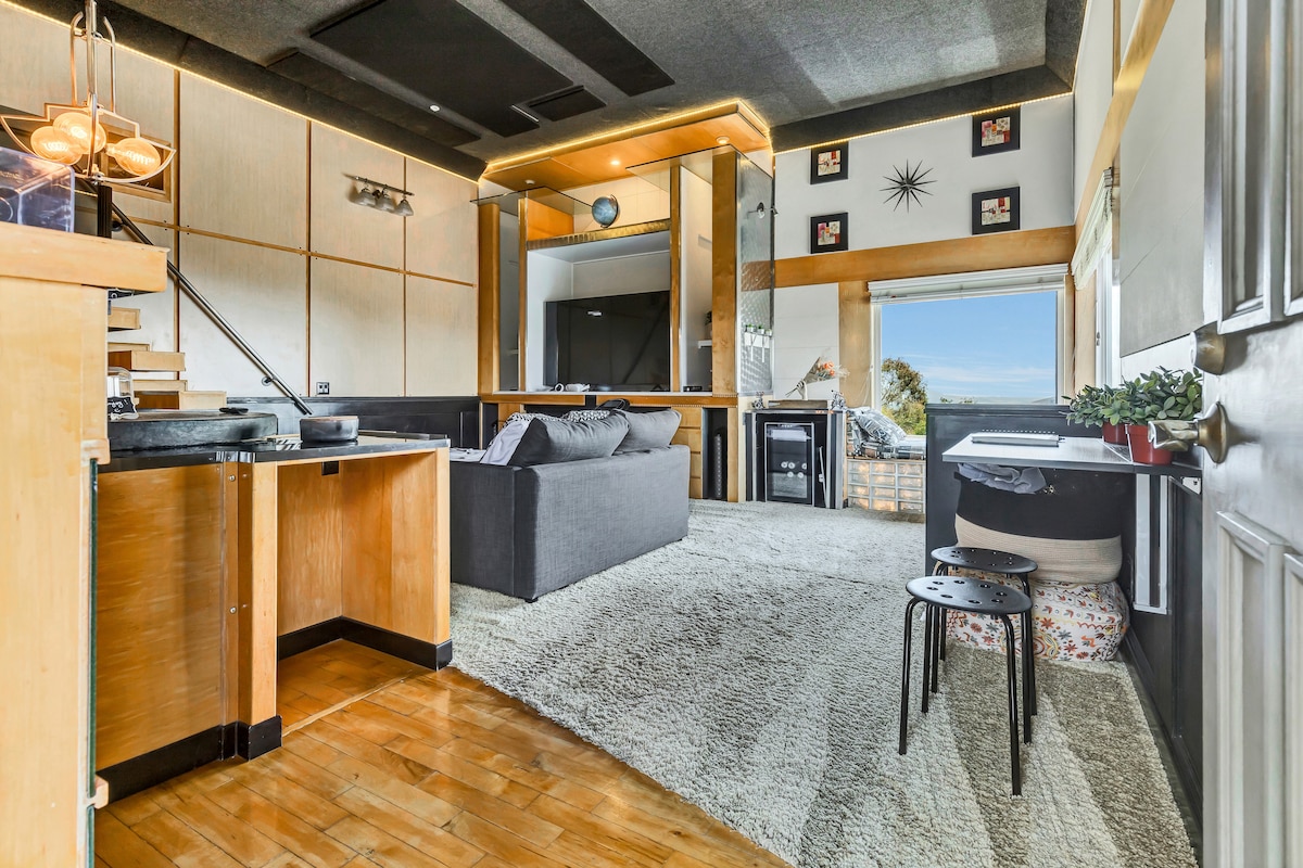 A modern loft space features warm wood flooring and a plush area rug. A gray sofa faces a sleek entertainment center. A desk and stool are situated by a window, offering views of the surrounding landscape. Natural light fills the room through large windows.