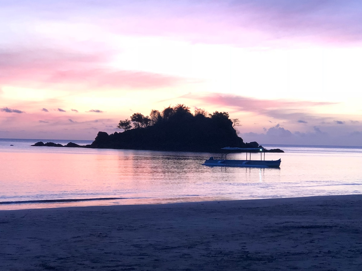 A tranquil scene of an island outlined by soft hues of pink and purple at sunset. Calm waters reflect the colorful sky, with a small boat anchored nearby. The tranquil beach scene showcases the serene ambiance of the coastal environment.