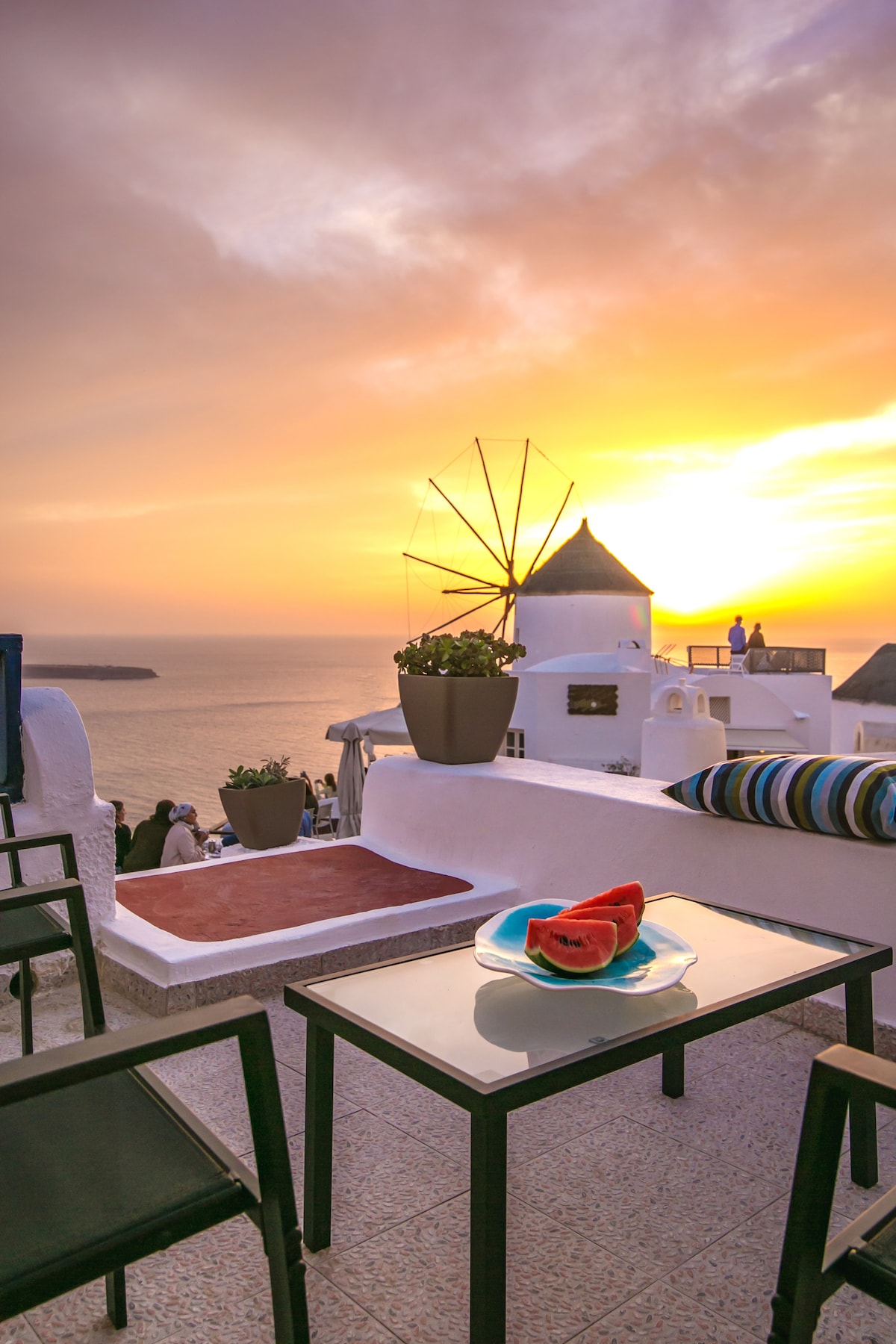 A private balcony is displayed with a dining table and chairs, facing a stunning sunset over the Aegean Sea. A plate with watermelon sits on the table, complemented by a potted plant and the silhouette of a nearby windmill.