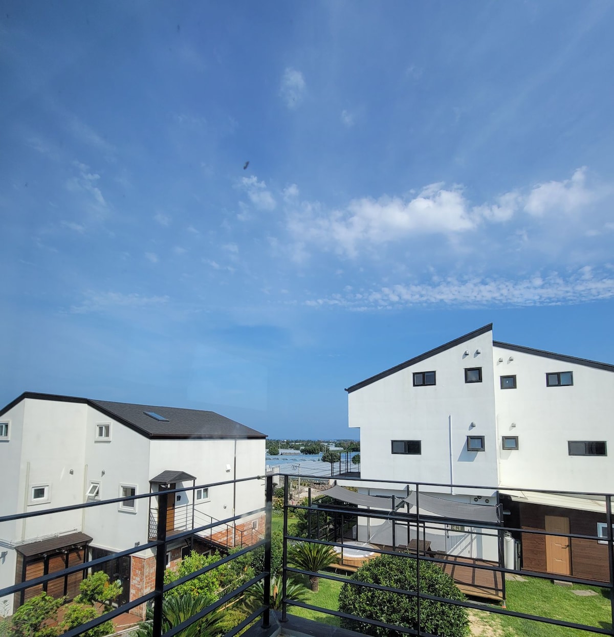 A clear blue sky fills the image, showcasing a peaceful neighborhood scene. Modern buildings with flat roofs and various window styles are visible, along with well-maintained greenery and outdoor spaces. The distance reveals a calm landscape, suggesting proximity to coastal views.