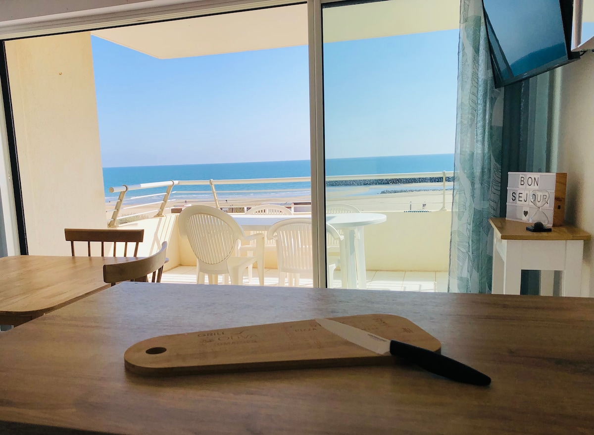 A spacious table is set in the foreground, featuring a wooden cutting board and knife. Large glass doors reveal a sunlit balcony with white chairs, offering a panoramic view of the calm sea and shoreline beyond.