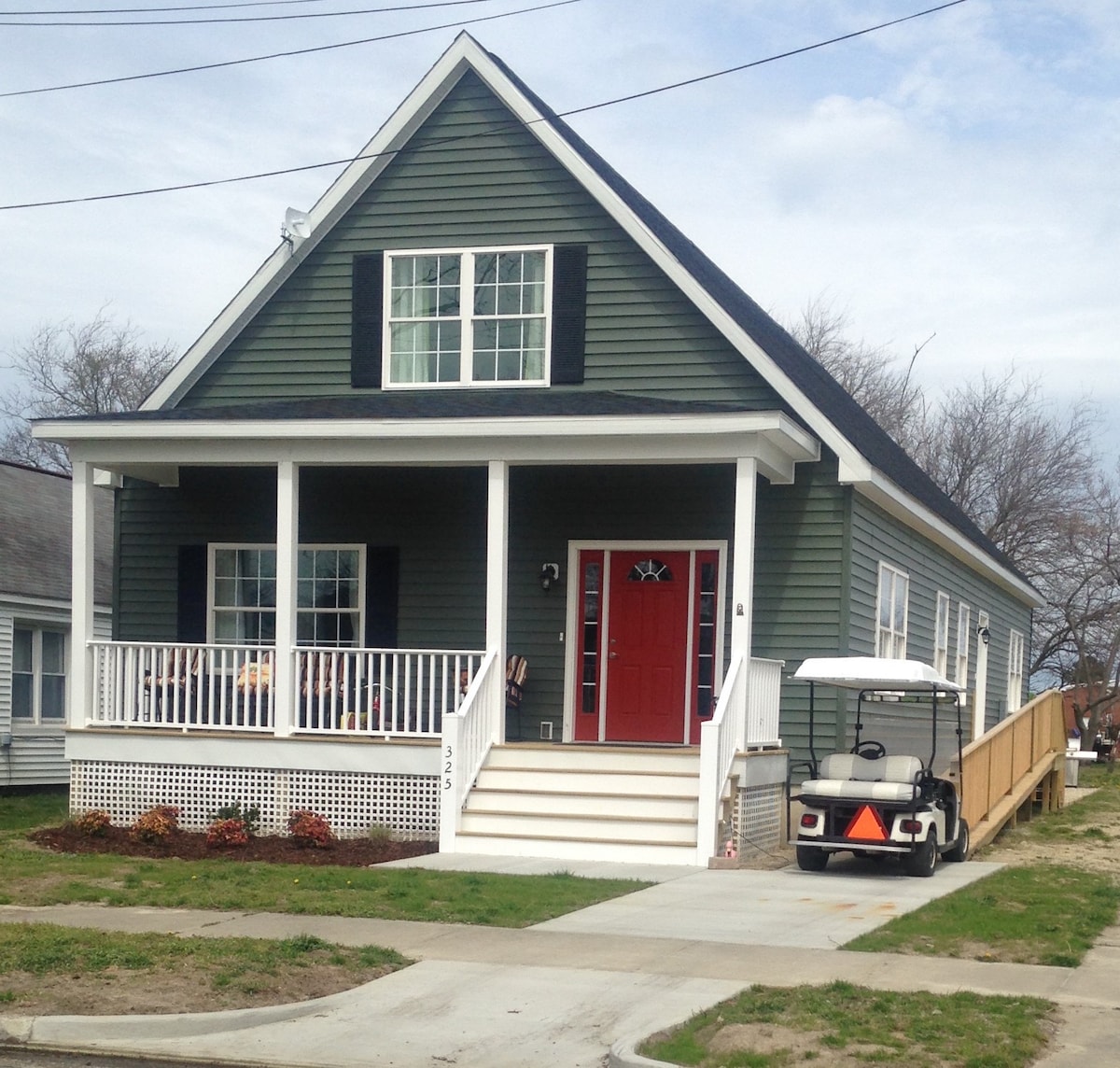 A green, two-story house with a prominent red door and white trim is visible. A spacious front porch features white railings, while a golf cart is parked nearby. A side door ramp enhances accessibility, and well-kept landscaping adds to the home’s welcoming appearance.