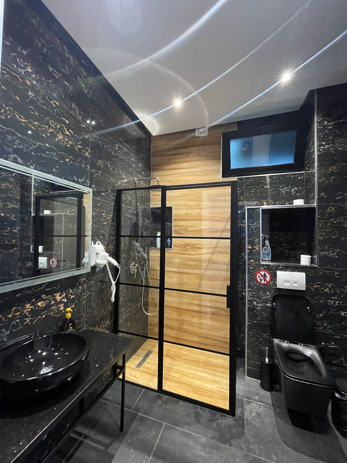 The bathroom features a modern design with dark stone walls and a sleek vanity with a round black sink. A spacious, glass-enclosed shower is visible, showcasing a wooden interior. Soft lighting from recessed fixtures enhances the overall ambiance, while a contemporary toilet is positioned nearby.